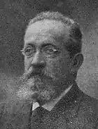 Une photographie en noir et blanc, légèrement floue, d'Ettore Molinari, un homme à la chevelure fuyante, aux lunettes étroites, à la moustache et à la barbe fournies.