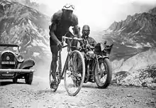 Photographie en noir et blanc d'un cycliste gravissant un col, suivi par une voiture et une moto.