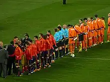 Des joueurs de football habillés en rouge et en orange se tiennent alignés, entourés d'arbitres et de cadreurs.