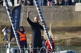 Debout à l'avant du bateau, le navigateur lève le poing. Près de lui, une fillette.