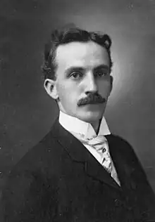 Homme en costume de trois-quarts face, cheveux courts de couleur sombre et moustache.