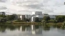 Bâtiment blanc, moderne, avec de grandes baies vitrées, se reflètant dans l’eau d’un lac.
