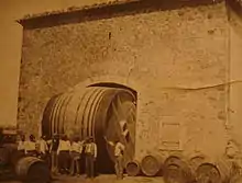 La photographie en noir et blanc jaunie par le temps indique une image issue de la fin du XIXe siècle ou du début du XXe siècle. Elle montre des barriques entreposées dans la cour d'un chai et huit ouvriers qui posent devant un foudre qui passe juste sous la voute du portail d'entrée de la cave.