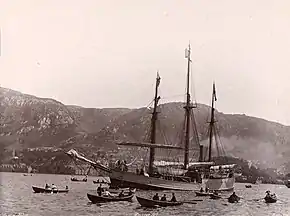 Un trois-mâts, propulsé à la vapeur, avance dans un bras de mer entouré de nombreuses barques à rame. À l'arrière-plan se profile une ligne de collines, avec quelques immeubles faiblement visibles au bord de l'eau.