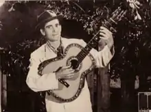 Photographie en noir et blanc d'un homme qui joue de la guitare. Il est vêtu de blanc et porte un chapeau. Derrière lui pousse des arbres.