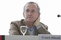 Photographie montrant Franck Riboud, cheveux gris et portant une paire de lunettes discrète, qui se tinet assis, sur la table devant lui, un verre vide et un écriteau où l'on distingue son nom