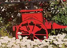 Photographie d'une charrette rouge et fleurie datant 1930 selon l'inscription écrite en noir, entourée de géraniums et de plantes d'exposition.