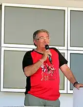 Photo en couleur d'un homme, debout, parlant devant un micro qu'il tient de la main droite.
