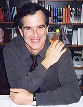 photo en couleur d'un homme assis derrière une table et devant une bibliothèque ayant la main droite sur l'épaule gauche et la main gauche tenant son coude droit