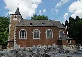 Édifice religieux en brique de style néo-roman. Au premier plan, un cimetière, en arrière plan, une forêt.