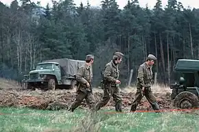 Trois hommes en uniforme et armés de fusils d'assauts marchent en colonne devant une bande de terre labourée. Deux camions se trouvent à l'arrière-plan devant une foret dense.