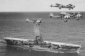 Photo en noir et blanc d'un porte-avions en mer, survolé par plusieurs biplans.