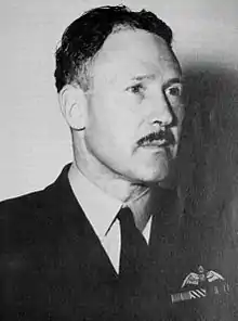 Portrait d'un homme moustachu en uniforme militaire sombre avec des ailes de pilote sur la poche gauche.