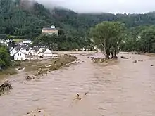Rivière en crue, au creux d'une vallée et bordée de maisons inondées ; en arrière-plan, une colline couverte de forêt.