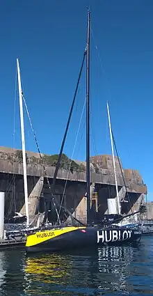 Vue de trois quarts avant, en entier, d'un voilier noir et jaune. En arrière-plan la base de sous-marins de Lorient.