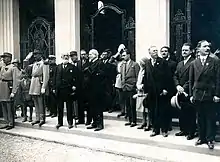 Photographie noir et blanc. Sur un perron une assemblée de personnages, du début du XXe siècle, en tenue de ville pose.