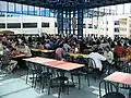 Grande et haute salle vitrée, à l’étage, tables de couleurs différentes selon les rangées et chaises.