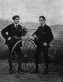 Photographie en noir et blanc montrant deux hommes se tenant debout de part et d'autre d'un vélocipède.