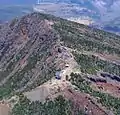 Photo couleur montrant le bord du cratère d'un volcan vu du ciel. Les pentes sont boisées et exposent des bandes de terre à nu par endroits. Trois bâtiments de petite taille sont visibles sur l'arête du cratère.