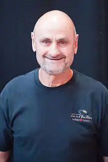 Photographie couleur d’un homme, de tronc, chauve, souriant, avec une barbe de 3 jours blanche et un t-shirt bleu marine