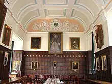 Un hall lambrissé de bois, avec un grand portrait d'Elizabeth I au milieu du mur du fond, d'autres portraits sur les murs ; un plafond de plâtre blanc à décor bleu clair