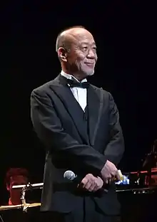 Un homme asiatique âgé se tenant debout, en smocking noir.