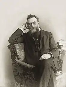 Homme barbu, assis sur un fauteuil et s'appuyant sur son coude, de trois quarts, avec quelques livres en fond.