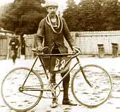 Photographie en noir et blanc d'un homme moustachu tenant son vélo, avec un tissu portant le no 28 au milieu du cadre.