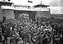 Photographie de déportés saluant un blindé devant une massive porte en pierre au-dessus de laquelle est déployée une banderole.