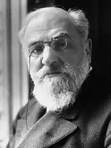 Photo en noir et blanc d’un homme aux cheveux et barbe blancs, portant des lorgnons et une veste épaisse