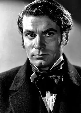 Portrait en noir et blanc d'un homme au regard dramatique. Il fait la moue et porte un manteau ainsi qu'un nœud papillon sombre.