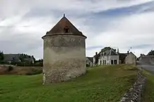 Photographie en couleurs d'un bâtiment circulaire à toiture pentue de tuiles s'élevant au milieu d'un pré, un muret en pierre visible sur la droite, des constructions anciennes et des maisons apparaissant en arrière-plan