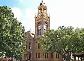 Vue partielle d'un bâtiment à deux étages, surmonté d'un clocher avec horloges, masqué en partie par des arbres