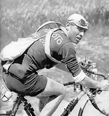 Photographie en noir et blanc d'un cycliste sur son vélo, portant un maillot avec l'inscription Automoto, une besace et des chambres à air en bandoulière, le regard porté vers la droite.
