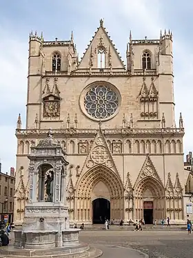 Vue en couleurs d’une cathédrale. Les toitures sont visibles derrière la façade. En arrière-plan, un cours d’eau.