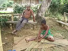 Une jeune malgache assise sur le sol, tresse des roseaux, pendant que son père est assis sur un banc à l'arrière.