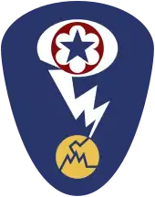Badge d'épaule ovale avec un fond bleu foncé. Au sommet se trouve une étoile bleue entourée d'un cercle rouge qui correspond à l'écusson de l'Army Service Forces. Ce dernier est entouré d'un ovale blanc représentant un nuage en champignon. Un éclair blanc sort du nuage et frappe un cercle jaune fissuré représentant un atome.