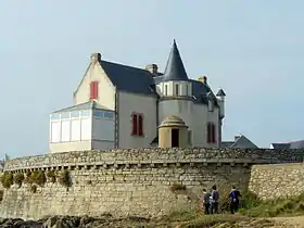 Vue d’un édifice massif avec tourelle pointue, retranché derrière une muraille de pierre.