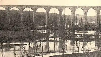 Photographie en noir et blanc d'un cours d'eau passant au-dessous d'un ouvrage d'art.