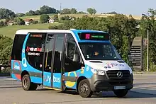 Photographie en couleurs d’un minibus bleu, blanc et noir circulant sur la route des Creuses devant un arrière-plan rural.
