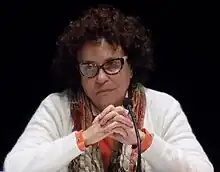 Photo en couleur d'une femme en buste, elle a les mains croisées et porte des lunettes