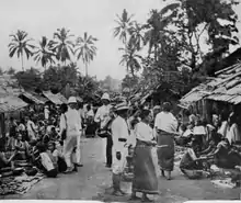 Un ensemble de cahutes en pailles, devant lesquelles sont assises des personnes autour de marchandises. Des personnes, debout, circulent entre les cahutes. On aperçoit deux hommes blancs, coiffés de casques coloniaux.