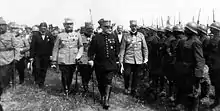 Photographie noir et blanc de trois hommes en uniforme paradant devant des troupes au garde à vous.