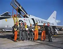 Sept hommes posant en combinaison de vol devant un avion militaire.