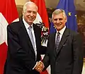 Photographie de deux hommes devant des drapeaux blancs, rouges, et bleus.