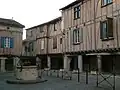 photo couleur de maisons sur une place à couverts. Le rez de chaussée comporte des portiques. Les étages sont à colombage garnis de brique foraine.