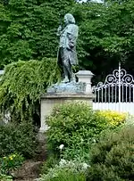 Statue en pied d'un homme en costume dix-huitième siècle, entourée de verdure.