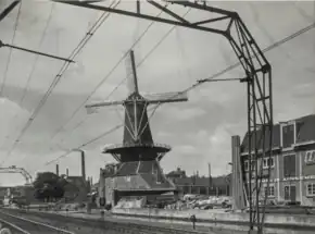 Photographie en noir et blanc d'un caténaire et des rails de chemin de fer traversant un paysage urbain, un moulin à vent et des bâtiments se dressant à droite de la ligne ferroviaire.