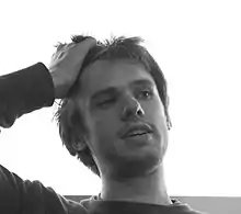Photographie en noir et blanc d'un homme en gros plan, blanc, brun, vu de face, regardant sur sa gauche, passant sa main droite dans ses cheveux.