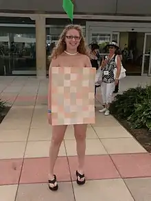 Femme portant une petite pancarte avec un patchwork de couleurs sur un ton rose en guise de robe. Portant seulement ce petit rectangle au niveau du buste, la personne semble nue.
